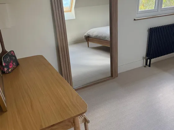Loft bedroom with an oak desk and stool, a tall arched oak-framed mirror reflecting a bed and roof window, a black column radiator and herringbone carpet