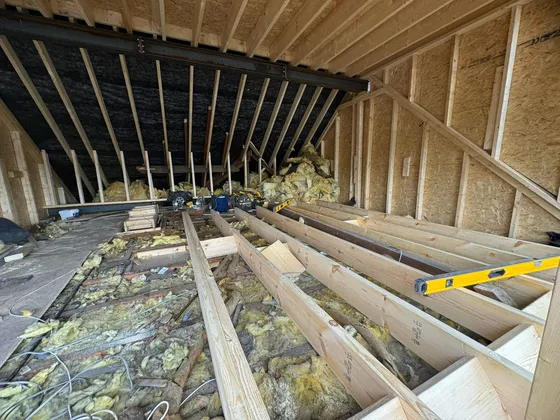 New timber floor joists laid across a loft over the old insulation, beneath a steel beam and rafters, with OSB-sheathed stud walls