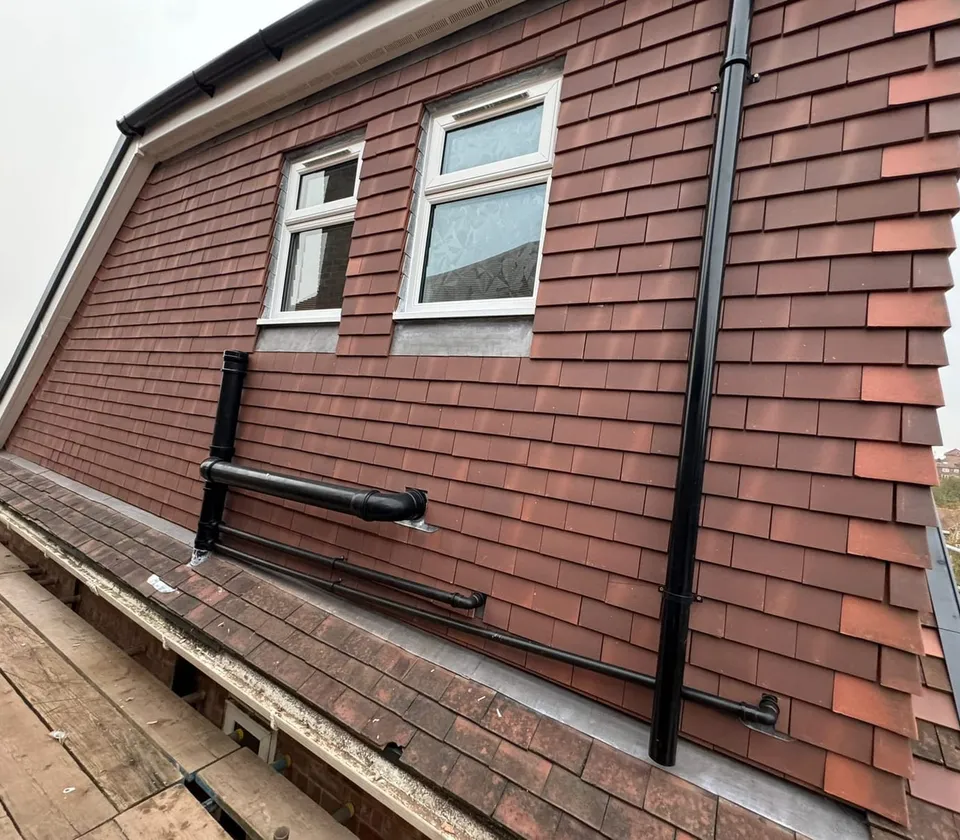 Large wall of red clay hanging tiles with two white windows, a black soil pipe and waste pipes, above scaffold boards