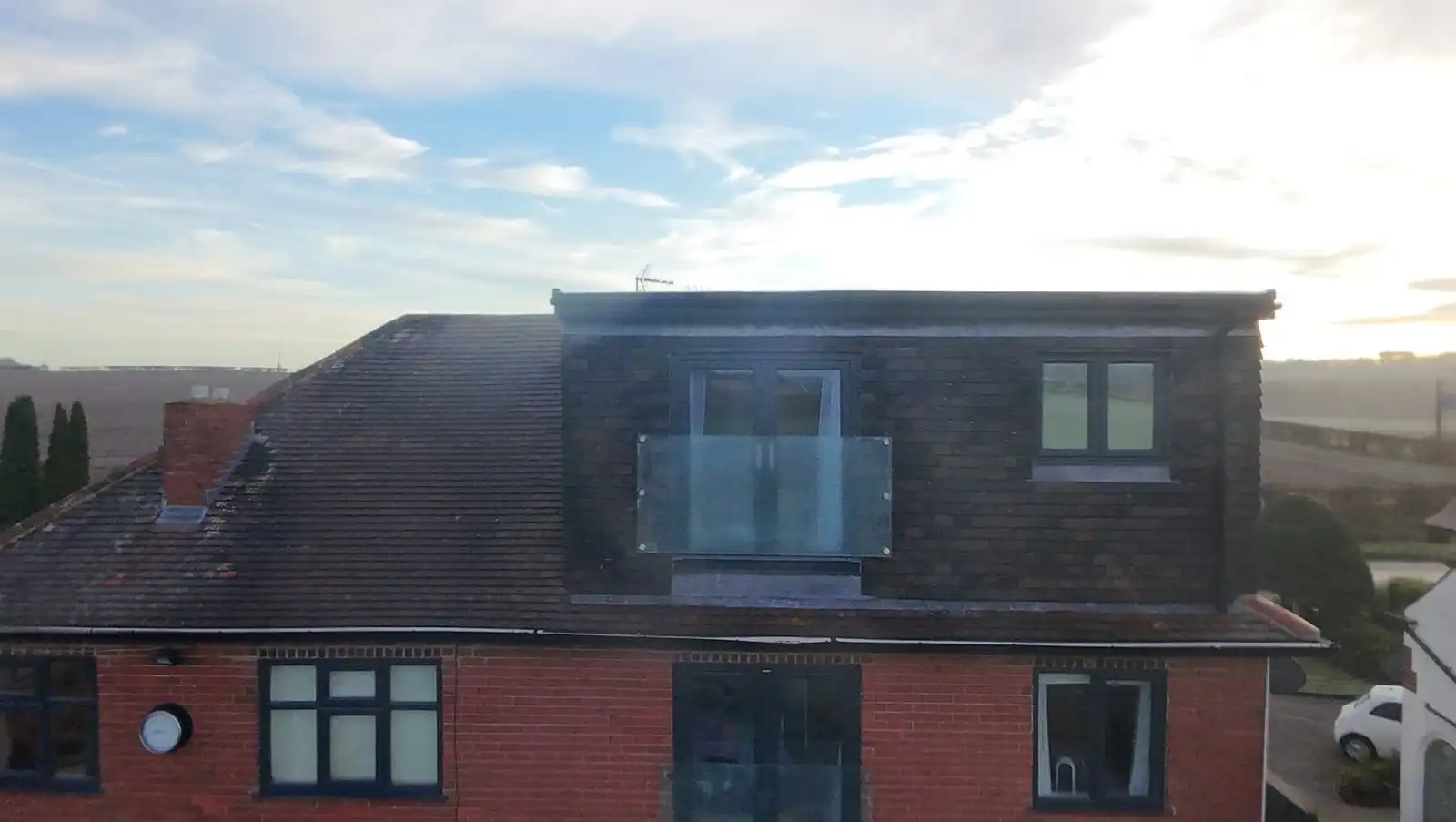 Aerial rear view of a flat-roofed dormer clad in dark hanging tiles, with a glass Juliet balcony and a grey window above red brickwork