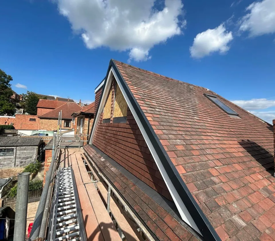 New gable with part-finished boarding and a roof window in the reclaimed clay-tiled roof slope, with scaffolding and a ladder along the side under a blue sky