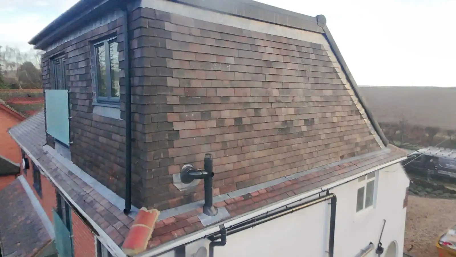 Close aerial view of the side of a dormer hung with multi-tone clay tiles, with a flue outlet and a glimpse of the glass balustrade on the front