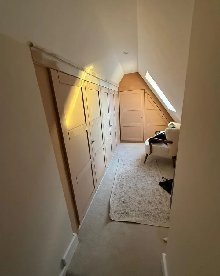 Narrow loft room with a run of fitted shaker-style wardrobes along the eaves, a roof window, an armchair at the far end and a long rug
