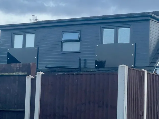 Flat-roofed dormer clad in grey boarding with three windows, two fitted with dark glass Juliet balustrades, seen over a garden fence